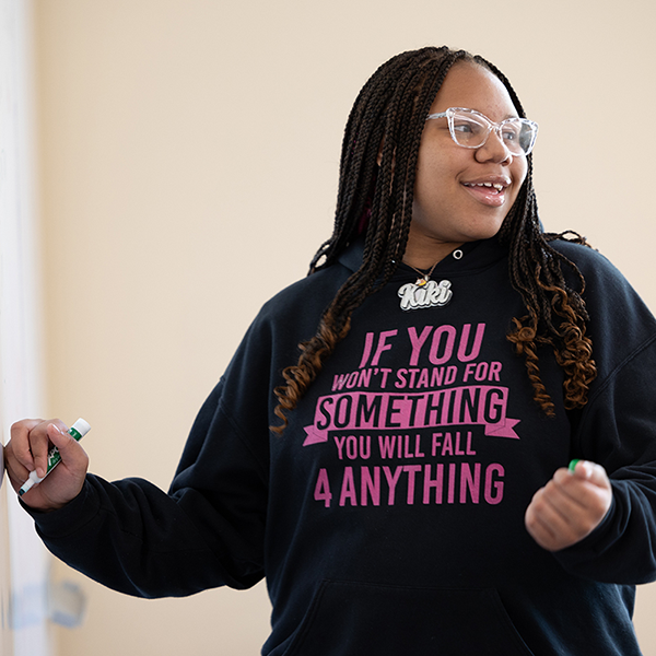 A Young Writers Institute participant writes on a whiteboard and smiles. She is wearing a black hoodie that reads in pink text: “If you won’t stand for something, you will fall 4 anything.”