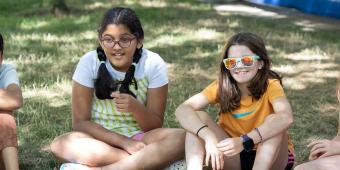 Girl with glasses sitting next to girl with sunglasses outside on the grass.